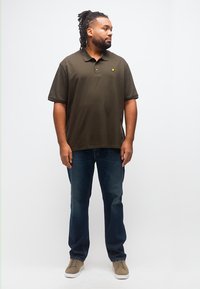 Άνδρας με ντραντλόκς, ντυμένος με ελιά πόλο πουκάμισο, σκούρο μπλε τζιν και μπεζ αθλητικά παπούτσια, στέκεται μπροστά σε λευκό φόντο, κοιτάζοντας προς τα δεξιά.