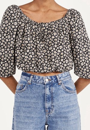 Blusa negra corta con un patrón floral que presenta margaritas blancas y amarillas, mangas abullonadas y un escote fruncido. Combinada con pantalones vaqueros azul de cintura alta.