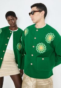 Grønne, broderte jakker med blomsterdesign, gullknapper og frontlommer. Modellen til venstre i en hvit topp og mønstrete skjørt.