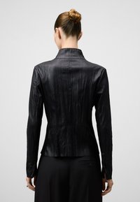 Giacca nera effetto tessuto con maniche lunghe, colletto alto e vita aderente. Il materiale sembra liscio con pieghe verticali.