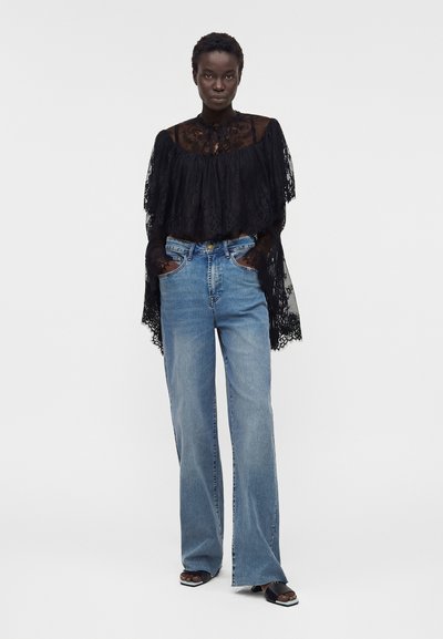 Γυναίκα στέκεται με τα χέρια στις τσέπες, φοράει μαύρο διαφανές δαντελένιο τοπ, μπλε φαρδιές τζιν και μαύρα σανδάλια με ανοιχτά δάχτυλα.