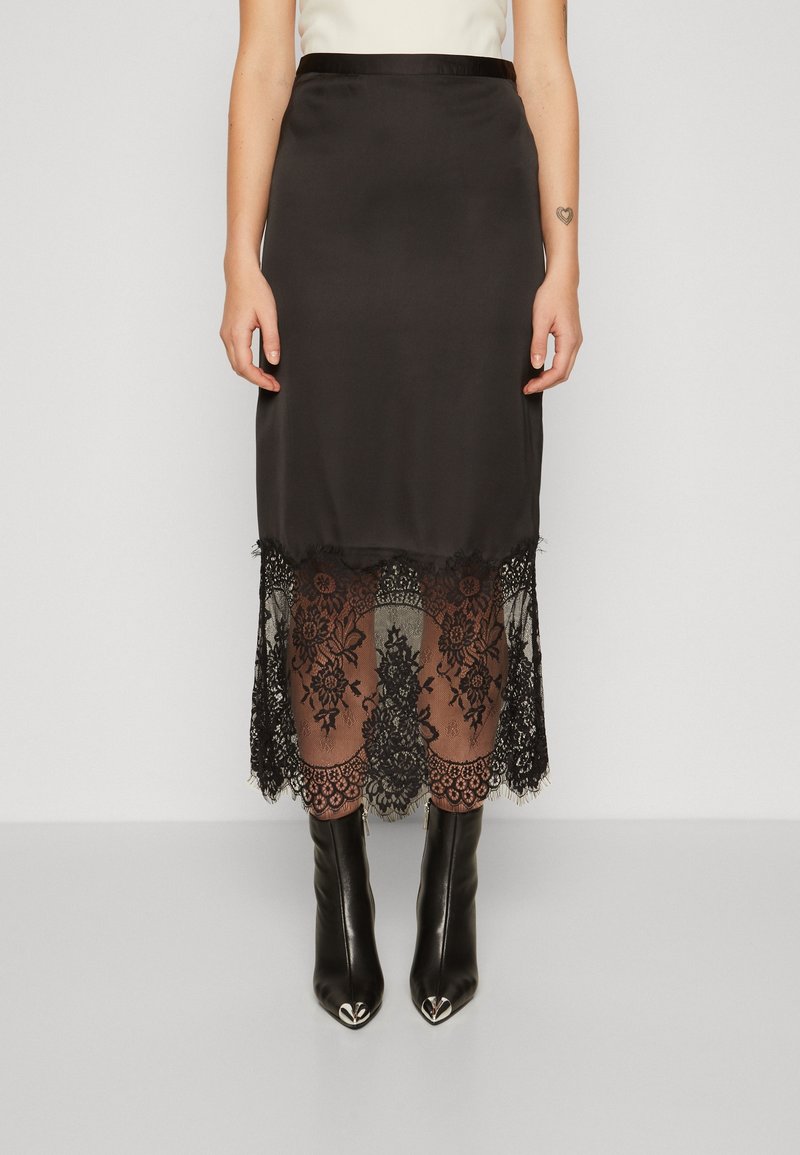 Femme portant une jupe en satin noire avec un ourlet en dentelle noire transparente, associée à des bottines noires à bout pointu avec des embouts métalliques.