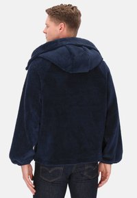 Granatowa, przytulna kurtka polarowa z dużym kapturem, luźnymi rękawami i miękką teksturą, noszona z ciemnymi spodniami dżinsowymi.