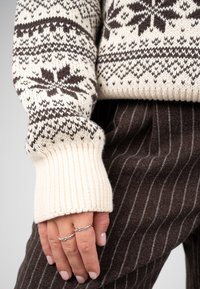 Main avec deux bagues en argent portant un pull en maille crème et marron à motifs et un pantalon marron foncé à fines rayures.