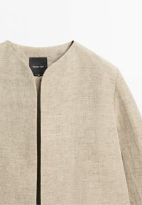 Beżowa lniana kurtka bez kołnierza i z otwartym przodem, z czarną metką Massimo Dutti wewnątrz dekoltu.