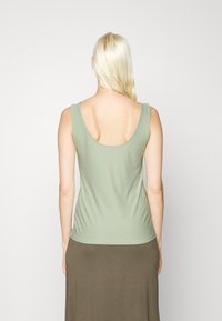 Top sem mangas verde claro com decote arredondado nas costas, feito de tecido macio, usado sobre uma saia longa castanha. Textura suave, design simples.