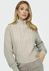 Hellgrauer Strickpullover mit hohem Kragen, der auf der Vorderseite Zopfstrickmuster und gerippte Bündchen sowie einen Saum aufweist. Reißverschlussdetail am Hals.