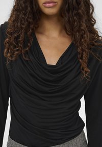 Melna garām piedurknēm virsdrēbe ar plūstošu apkakli un smalku tekstūru. Iezīmējas ar savilkta auduma un pieguļošu siluetu.