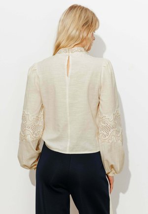 Femme aux cheveux blonds portant un chemisier crème orné de dentelle avec des manches bouffantes et un pantalon noir, vue de dos sur un fond blanc.