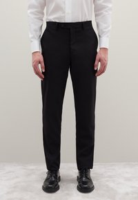 Zwarte, op maat gemaakte broek met een rechte pasvorm, voorzien van een knoopsluiting en zijzakken, gecombineerd met glanzende zwarte veterschoenen.