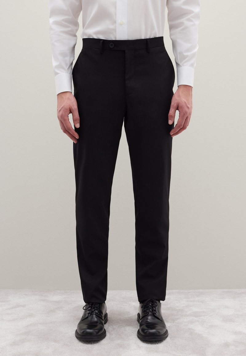 Zwarte, op maat gemaakte broek met een rechte pasvorm, voorzien van een knoopsluiting en zijzakken, gecombineerd met glanzende zwarte veterschoenen.