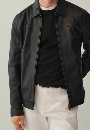 Homme portant une veste en cuir noire sur un pull noir et un pantalon blanc, une main reposant sur le côté et l'autre derrière le dos.