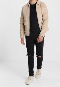 Veste en velours côtelé beige avec deux poches avant, portée sur un t-shirt noir, associée à un jean noir déchiré et des baskets beige.
