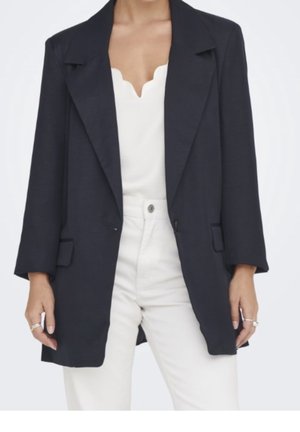 Femme portant un blazer noir oversize sur un haut blanc à col en V festonné et un pantalon blanc taille haute, avec des bagues aux deux mains.