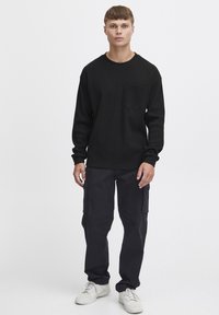 Black waffle-knit sweatshirt with long sleeves and a chest pocket, paired with dark cargo pants and white sneakers. Simple, functional design.