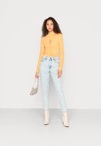 Top laranja de mangas longas com um recorte em forma de olho-chave, combinado com jeans de lavagem clara. Segura uma pequena bolsa xadrez; usa botins creme até ao tornozelo.