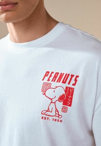 White cotton t-shirt featuring a red graphic print of Snoopy with text "PEANUTS" and "EST. 1950" in a cartoon style.