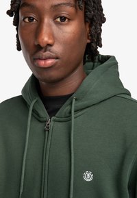 Sudadera verde con cremallera hecha de tela suave, que cuenta con una cremallera frontal, capucha con cordón y un pequeño logo blanco en el pecho.