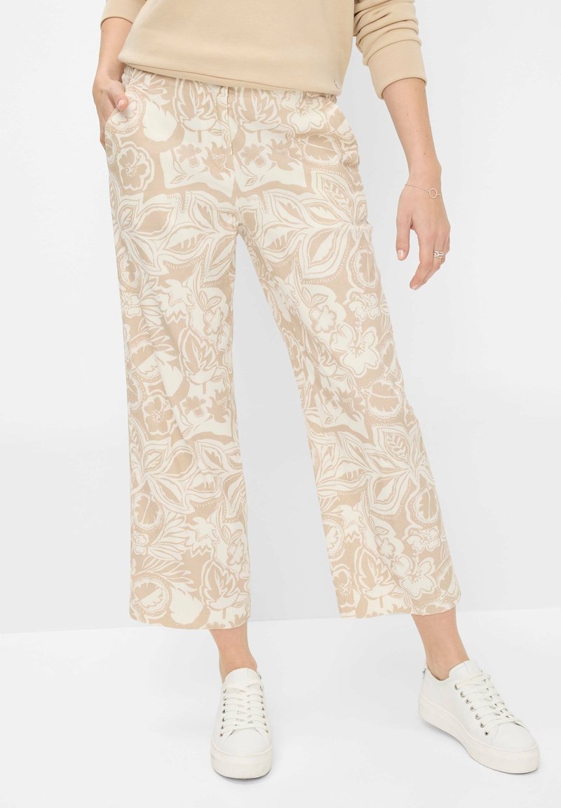 Femme portant un pantalon beige à jambes larges avec des motifs floraux blancs, un haut à manches longues beige et des baskets blanches, debout avec une main dans la poche.