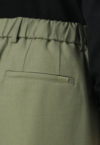 Light green trousers with an elastic waistband, featuring a back pocket and a small logo tag. Fabric has a subtle texture.