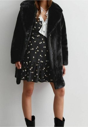 Femme portant un manteau en fausse fourrure noire sur une robe noire à fleurs avec un détail de nœud blanc, associée à des bottines noires.