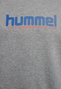 Grå sweatshirt med blå och röda "hummel"-logotext. Materialet verkar mjukt med en melerad textur, vilket betonar en avslappnad design.