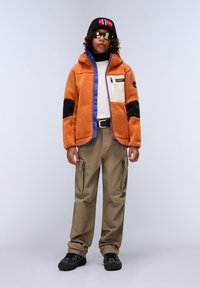 Oransje fleecejakke med blå og svart detaljer, kremfarget brystlomme; beige cargo-bukser; svarte sneakers; svart lue med flaggdetalj; solbriller.