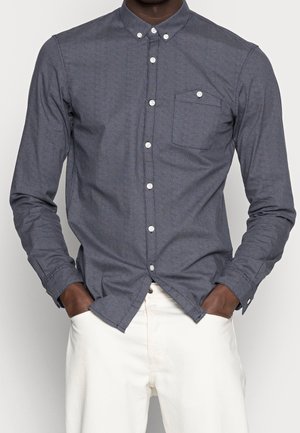 Hombre vestido con una camisa de botones de color gris oscuro con un bolsillo en el pecho, con las manos en los bolsillos, vestido con pantalones blancos, de pie frente a un fondo liso.