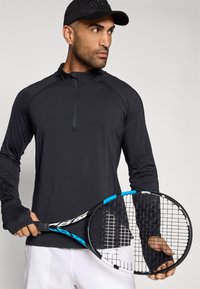 Μαύρο αθλητικό μπλουζάκι με μακρύ μανίκι, μισό φερμουάρ στον γιακά και τρύπες για τα δάχτυλα. Κρατάει μια ρακέτα τένις με μπλε λεπτομέρεια στη λαβή.