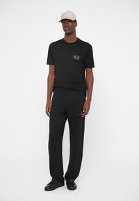 T-shirt preta de algodão com patch de logótipo, combinada com calças de fato de treino pretas. Boné bege notável completa o conjunto, apresentando um design estruturado e aba plana.