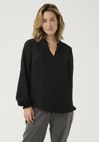 Black long-sleeve blouse with a V-neck and ruffled collar, made from textured fabric. Loose fit with gathered cuffs. Paired with gray trousers.