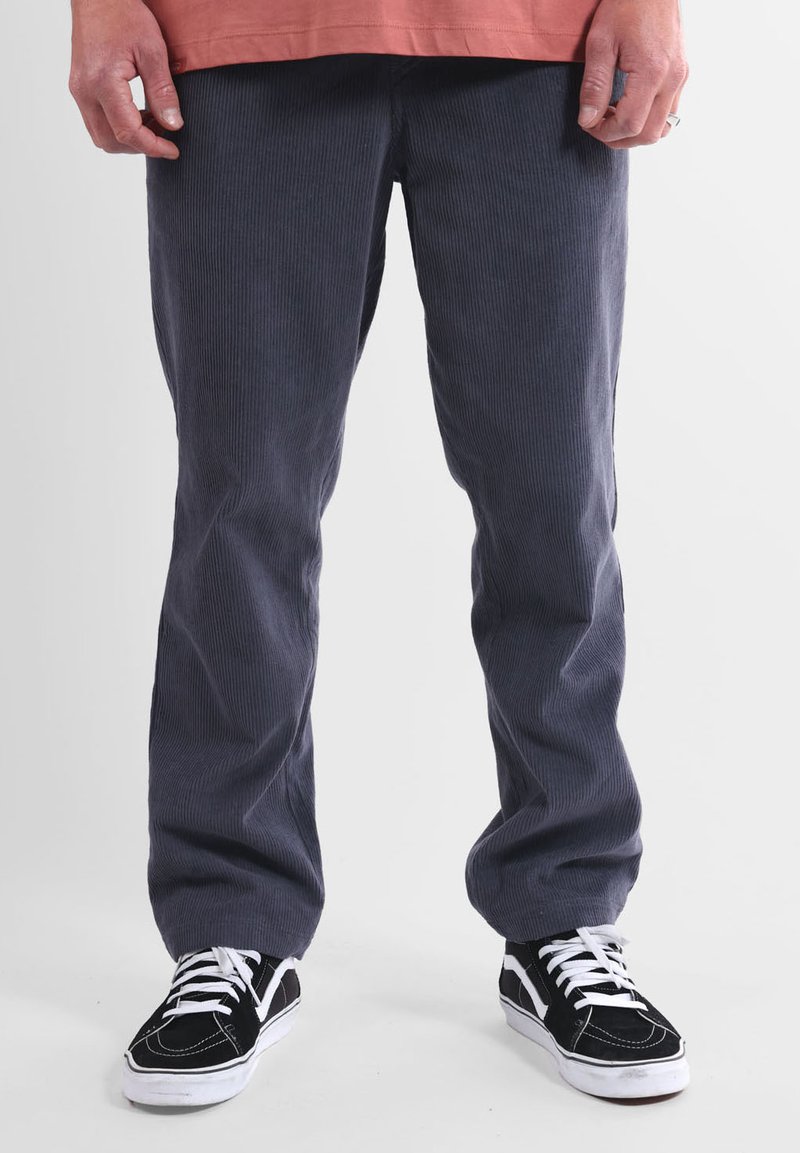Pantalon en velours côtelé gris avec une texture côtelée verticale, coupe droite, associé à des baskets noires avec des lacets blancs et des bandes latérales.