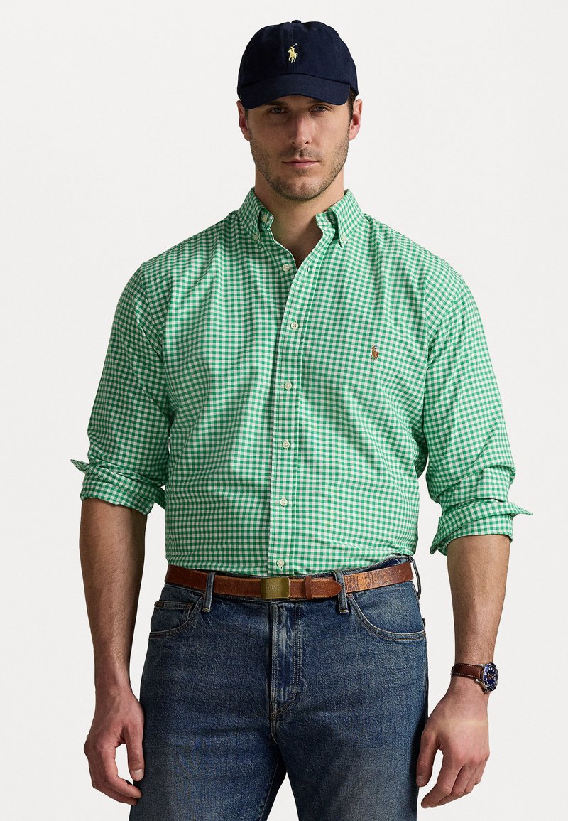 Chemise à carreaux verte et blanche à manches longues, accompagnée d'une casquette bleu foncé, d'une ceinture marron et d'un jean bleu. Montre-bracelet simple visible.