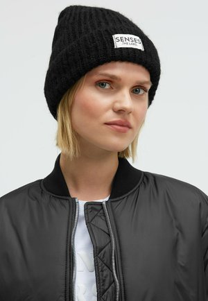 Black knitted beanie with a ribbed texture and a white sewn label reading "SENSES THE LABEL." Model wears a black bomber jacket over a white shirt.