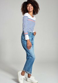 Witte blouse met lange mouwen en blauwe en rode strepen, gecombineerd met lichtblauwe high-waisted jeans en witte sneakers. Model staat met één hand op de heup.