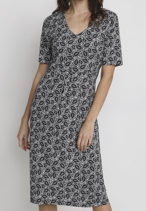 Femme portant une robe noire et blanche à motif paisley longueur genoux, manches courtes et col en V sur un fond uni.