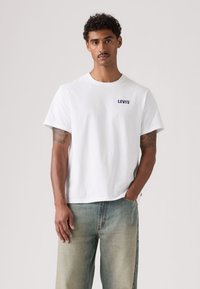 Λευκό βαμβακερό μπλουζάκι με κοντά μανίκια και στρογγυλή λαιμόκοψη· διαθέτει σκούρο μπλε λογότυπο "LEVI'S" στο στήθος· φοράται με ανοιχτό μπλε ξεθωριασμένο τζιν.