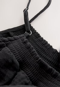 Tessuto nero strutturato con motivo a coste, caratterizzato da sottili spalline e accenti in metallo argentato. Vista ravvicinata che mette in risalto i dettagli delle cuciture.