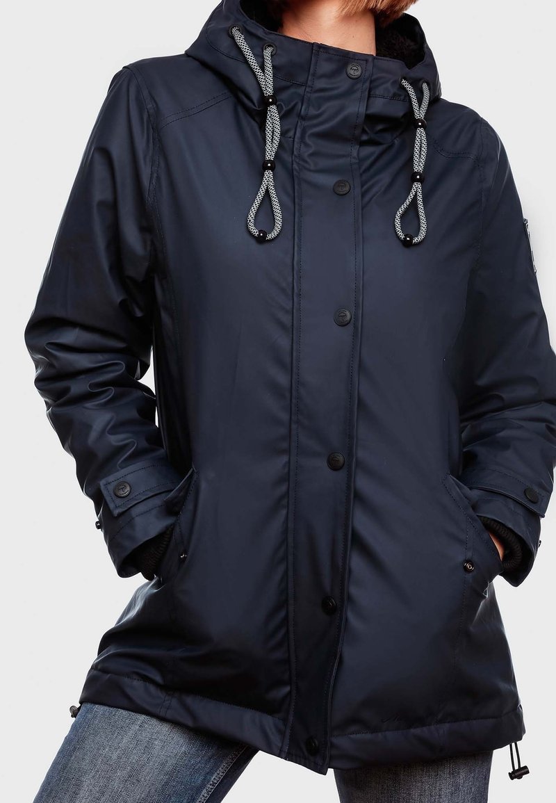 Chaqueta impermeable azul marino con capucha, que presenta un cordón regulable, bolsillos con cremallera y cierres de botón. Textura suave y corte casual.