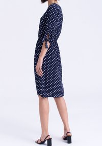 Námořnické šaty s puntíky, se třičtvrtečními rukávy, stahovaným pasem a mašlovými detaily. Hladká látka a délka až ke kolenům. Ve spojení s sandály na blokovém podpatku.