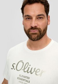 Biała koszulka z logo "s.Oliver" w kolorze złotym, okrągły dekolt, krótkie rękawy, widoczna tekstura materiału, częściowo pokazana twarz modela.