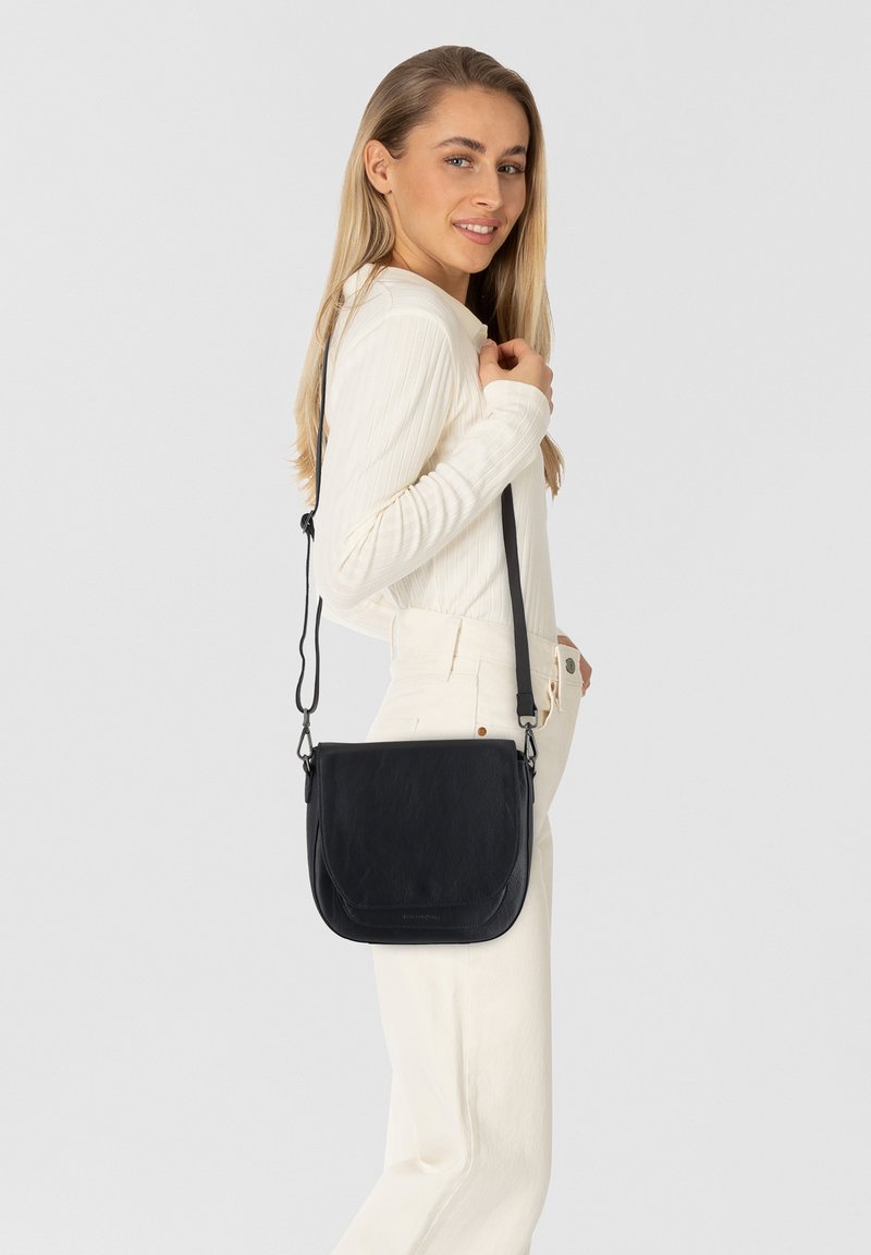 Giovane donna in abbigliamento crema sorride mentre porta una borsa a tracolla in pelle nera sulla spalla, su uno sfondo neutro.
