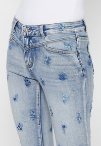 Ljusblå denimjeans med blomsterbroderi i olika blå nyanser. Har en klassisk femficksdesign och en silverfärgad knappknäppning.