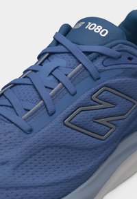 Κοντινό πλάνο ενός μπλε αθλητικού παπουτσιού New Balance 1080 με επάνω μέρος από δίχτυ, λεπτομερείς ραφές και επίπεδα κορδόνια δεμένα σε σταυρωτό σχέδιο.