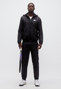 Νέος άνδρας με μαύρα αθλητικά ρούχα κρατώντας ρακέτα τένις, στέκεται μπροστά σε απλό λευκό φόντο.