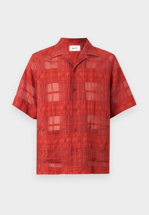Hombre con camisa roja de manga corta con textura, pantalones cortos negros hasta la rodilla y sandalias negras sin cierre, de pie con las manos en los bolsillos.