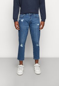 Jeans de denim azul claro desgastados com um corte cropped, apresentando múltiplos rasgos e bainhas desfiadas, combinados com ténis brancos.