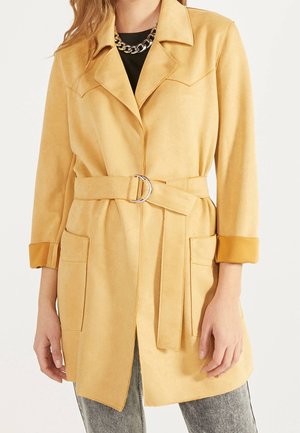 Femme portant un manteau ceinturé de couleur beige clair avec de grandes poches avant, des manches retroussées, un collier chaîne argenté et un jean gris délavé.