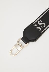 Cintura nera regolabile con hardware dorato e dettagli in pelle, caratterizzata da un design intrecciato e testo bianco lungo tutta la lunghezza.