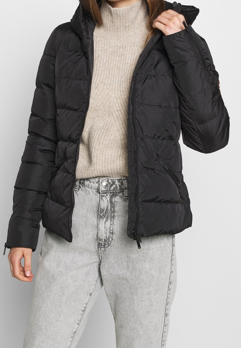 Black puffer jacket with a hood, quilted design, zippered front, and side pockets. Worn over a cream ribbed sweater and grey jeans.
